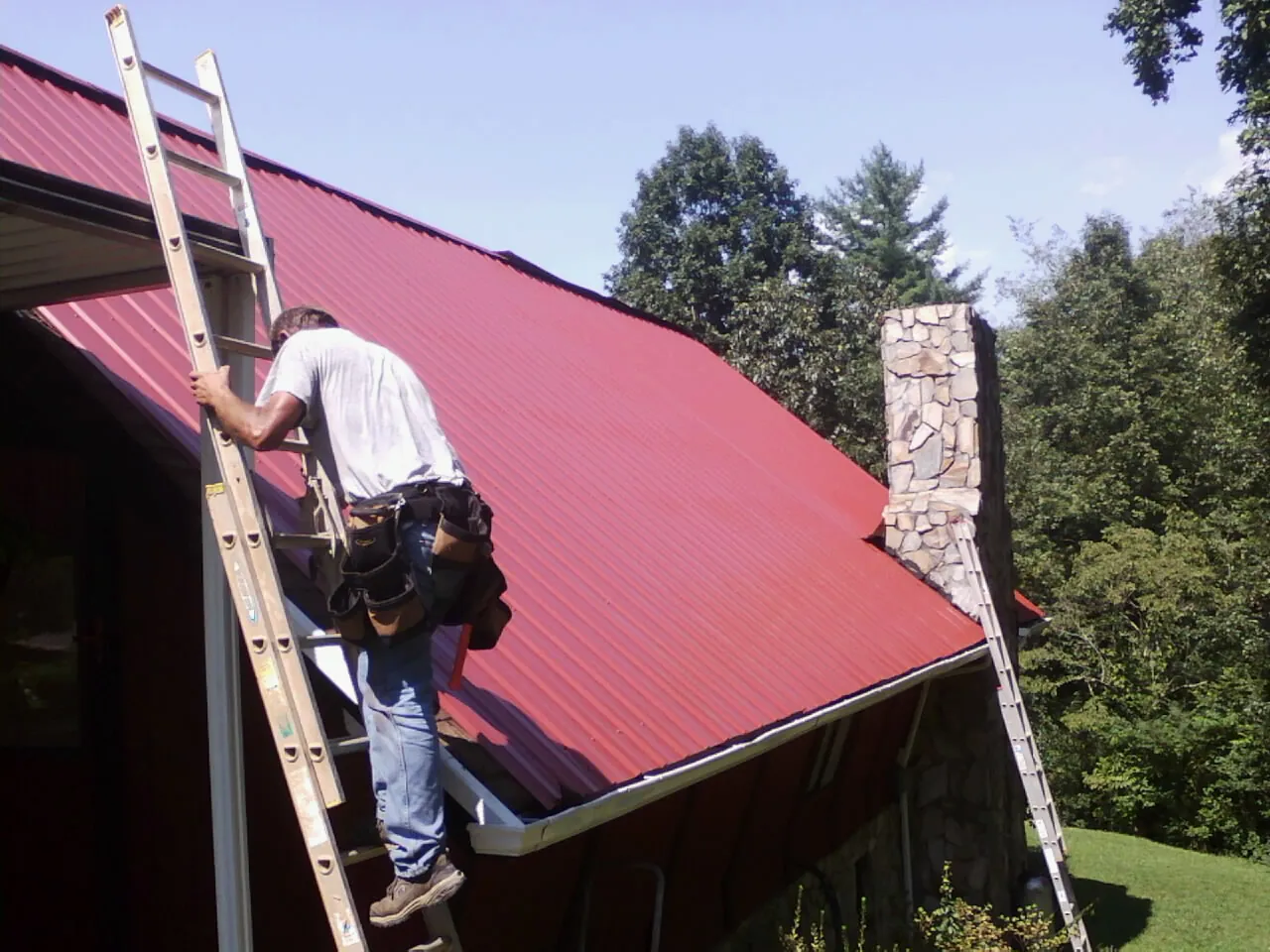 Roofer on a ladder beside a red metal roof and stone chimney — Soffit & Fascia Repair by Roofing Brothers in Greeleyville
